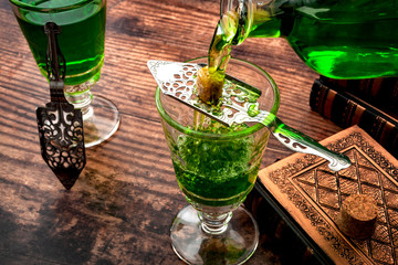 Alcoholic drink, creative stimulant and bohemian lifestyle concept theme with a vintage glass bottle pouring absinthe over a sugar cube in a stainless steel spoon next to books on a wooden table - obrazy, fototapety, plakaty
