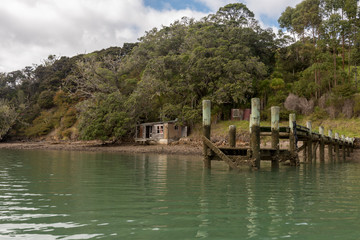 An old, wooden wharf and the ruin of an abandoned beach house on Motukauri Island in Whangaruru Harbour, Northland, New Zealand. Viewed from the sea.