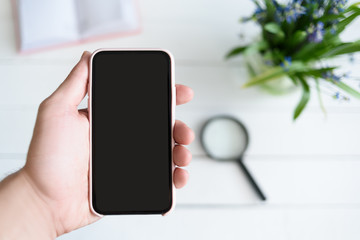 Male hand with a smartphone. Black blank screen. Table with notebook and flowers on background