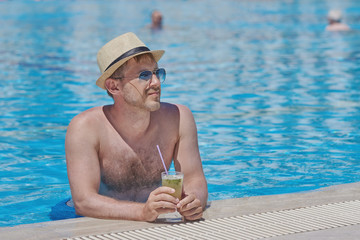 The portrait of a handsome man in a sun hat and sunglasses with the mojito cocktail in his hand. He is relaxing in the pool and enjoying his vacations.