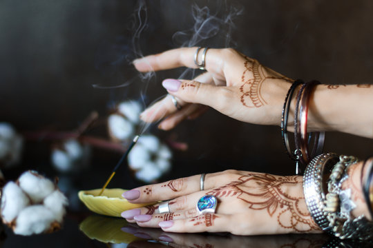 Closeup of female wrists painted with henna traditional Indian oriental mehndi ornaments. Hands dressed in bracelets and rings put aromatic stick in stand.  Branch with cotton flowers on background.