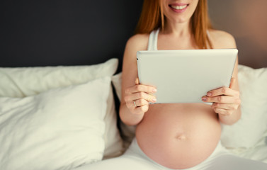 Technology and pregnancy. Young pregnant  woman using tablet computer sitting on a bed at home.