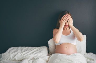 Tired stressed pregnant woman holding hand on face while sitting on a bed.