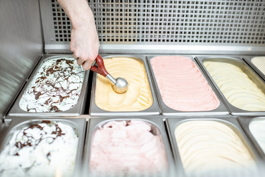 Metal trays full of colorful ice cream in the showcase refrigerator, salesman taking ice cream with scoop, close-up view