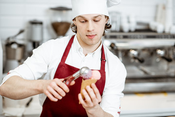 Young salesman in apron and hat making ice cream with waffle cone at the modern pastry shop