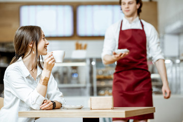 Handsome waiter in red apron serving sweet cake for a young woman customer at the pastry shop or cafe