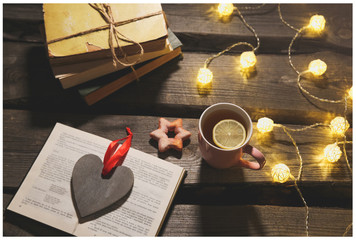 Holidays mood photo. Christmas lights and hot tea mug. Book for cosy evening. Sweet gingerbread and wooden heart on tray. Perfect winter flat lay. Hygge concept.