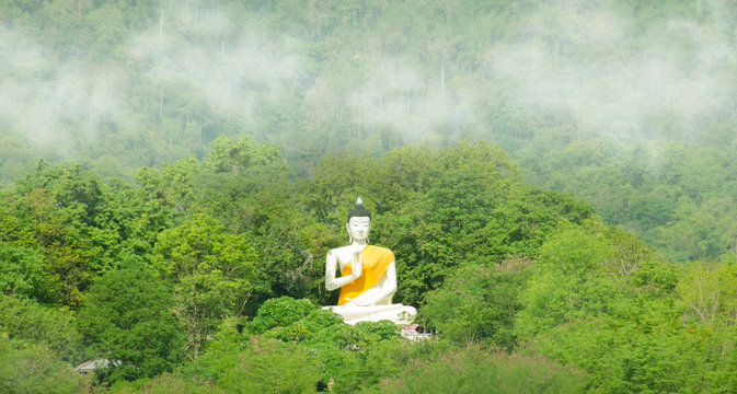 The large white Buddha statue is located on a mountain with green forests, very cloudy in Thailand.