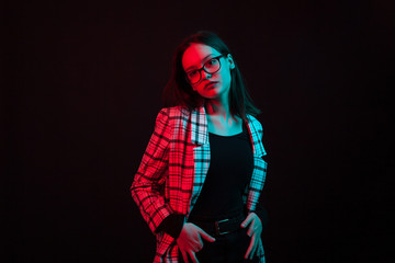 Portrait of cute young girl keeping her hands in pants pocket and looking at the camera in studio with colorful lighting