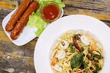 Top view seafood stir fried spicy spaghetti in white ceramic plate with smoked sausage on wooden table in the restaurant.