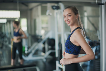 Fototapeta na wymiar Young sport Caucasian woman shot hair is measuring waist in gym near big mirror. Slim woman use measuring yellow tap and looking at camera in sport club. Woman felling happy and smiling.