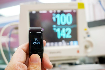 Hand holding black healthy smart band with numbers and icon of hearth rate and pulse display on screen with blurry of BP, vital sign and Heart rate monitor in hospital.