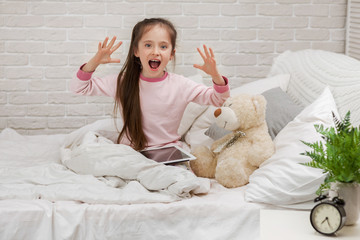 surprised little child girl lies in bed uses digital tablet. child playing on tablet pc.