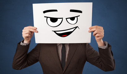 Casual person holding a paper in front of his face with drawn emoticon face 
