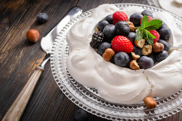 Pavlova cake with fresh summer berries and hazelnuts on glass standcake  on black wooden table.