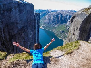 Obraz premium A girl lying at the edge of a steep mountain pretends to be flying. She spreads arms wide, in a gesture of freedom. In front of her stunning Lysefjorden shimmering with many shades of blue and green