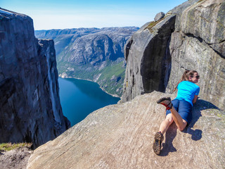 A girl lying at the edge of a steep mountain. She is shaking her legs, enjoying the herself and the view. In front of her stunning Lysefjorden shimmering with many shades of blue and green