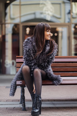 beautiful brunette with the long straight hair wearing in a grey fur coat, white shirt and black skirt siting  on the bench and smiles