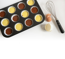 muffin baking tray filled with light and dark cake dough for cupcakes, whisk, eggshell and  paper cups, background fades to white, large copy space, high angle view from above