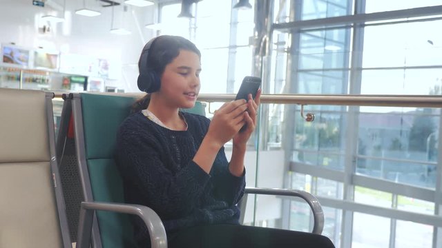 airport waiting hall room for a flight by plane. young happy teenage girl in headphones listening to the music on smartphone chatting communicates in the messenger. teenager girl in social networks