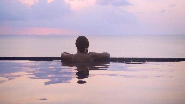 Woman relaxing at the edge of infinity swimming pool at resort. Young Female watching beautiful Sunset View, turns and looks into camera. She is enjoying vacation at resort by sea. Pink, orange violet