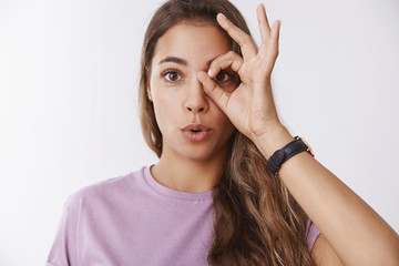Fototapeta premium Cute playful curious young female university student showing circle okay gesture on eye looking through amazed saying wow folding lips amused astonished, seeing incredible awesome product