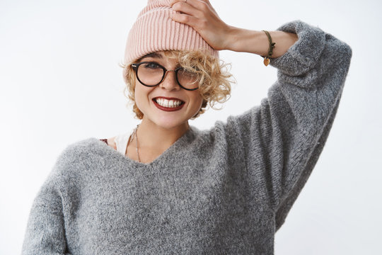 Getting all trendy this winter season. Portrait of charming blond european girlfriend in sweater and pink beanie pulling hat on forehead and smiling broadly being in good mood over white wall