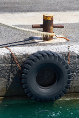 Old truck tire hanging on metal chains protecting shipps hulls and a mooring rope around a rusty harbor bollard at a port pier in Greece.