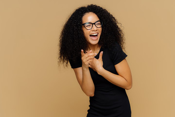Indoor shot of overjoyed female laughs out loudly, notices funny scene, points at camera, keeps...
