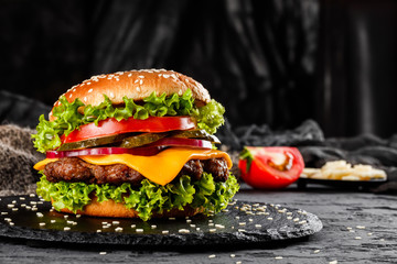 Beef burger with cheese, tomatoes, red onions, cucumber and lettuce on black slate over dark background. Unhealthy food - obrazy, fototapety, plakaty