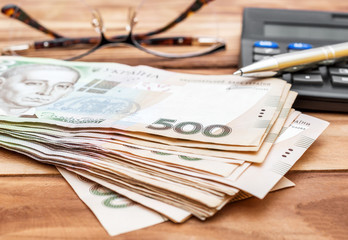 Heap of hryvnia bills with calculator and eyeglasses on wooden table. Close up.