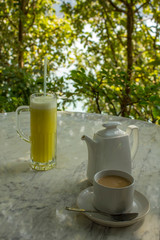 bright yellow drink in a glass beaker with a straw and a cup with a milk drink near a white ceramic teapot on a marble table on a blurred background of green trees