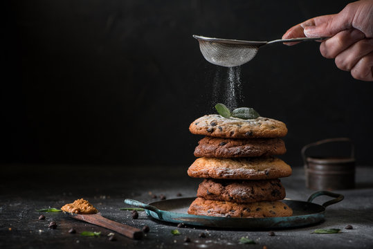 chocolate cookies with cereals and raisin in vintage baking pan. Woman's hand with sieve sprinkling sugar powder from above, dark plywood wall background