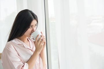 Side view portrait a pretty woman thinking looking holding a coffee mug beside a window at home