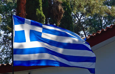 Large Greek flag flapping in a park in front of church roof with tiles.