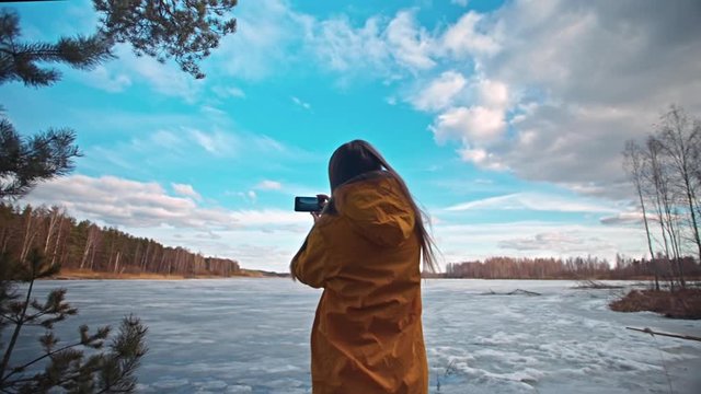 Woman with long hair in a yellow jacket takes pictures of a beautiful blue sky with clouds over a frozen forest lake in spring