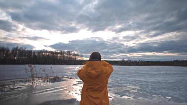 Woman in a yellow jacket stands on the bank of a frozen lake, takes a phone from her pocket and takes pictures of the cloudy sky and the forest on the other side of the lake, a spring trip, nature.