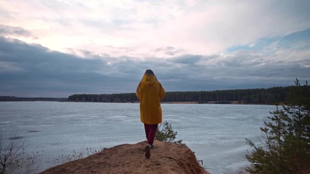 Woman in a yellow jacket walks to a cliff and takes pictures of a beautiful sky with clouds and a forest on the other side of a frozen lake on her smartphone. Spring trip, nature.