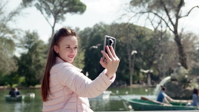 Pretty girl with long brown hair and adorable smile making selfie photos on the background of lake. Beautiful lady holding modern smartphone in her hands. Young model posing near the lake in the park