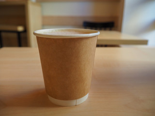 Cup of cappuccino in the paper takeaway cup on the wooden table in cafe close up