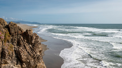 Spring afternoon at Ocean Beach in San Francisco.  High angle shot looking south from Cliff House.  Mild surf, people enjoying a relatively warm day.  Blue sky.  Rough cliffs foreground.