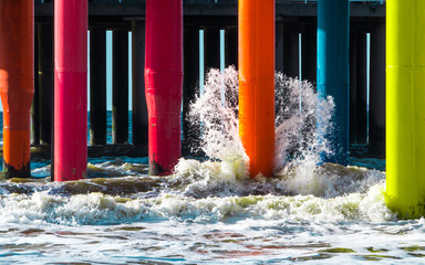 Wave brakes into orange colorful pier footing lit up with a sun with other rainbow color pier...