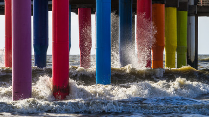 Wave brakes into colorful pier footings lit up with a sun and sparkling with other rainbow color...