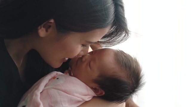 Beautiful Asian smiling mother is holding her newborn sleeping baby on chair next to window. She use her nose touching softly on baby nose. She loves her child so much. Love from mother concept.