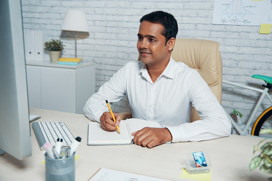Confident Indian businessman sitting at the table looking at computer monitor and making notes in his notepad at office