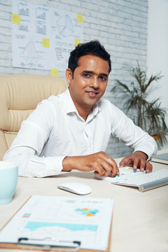 Portrait of Indian mature manager sitting at the table with computer and documents and smiling at camera at modern office