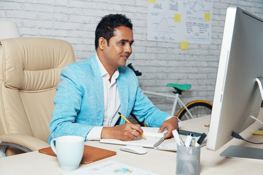 Indian mature businessman sitting at the table in stylish suit looking at computer monitor and writing business plan in his notepad at office