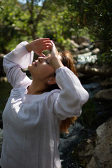 Latina woman holding her temples back with her hair in the sun in front of woods and a stream in the shade