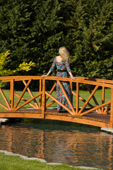 Beautiful long haired blonde woman stand on a wooden bridge over an artificial pond, in natural surround, green background.