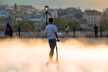 Boy on a scooter in a fog of sprayed water. Summer in old european sity